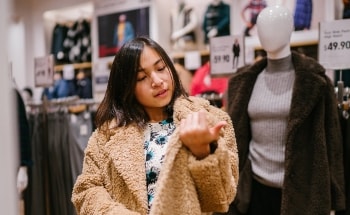 A young woman trying on a fuzzy sweater in a shop while looking at her nails